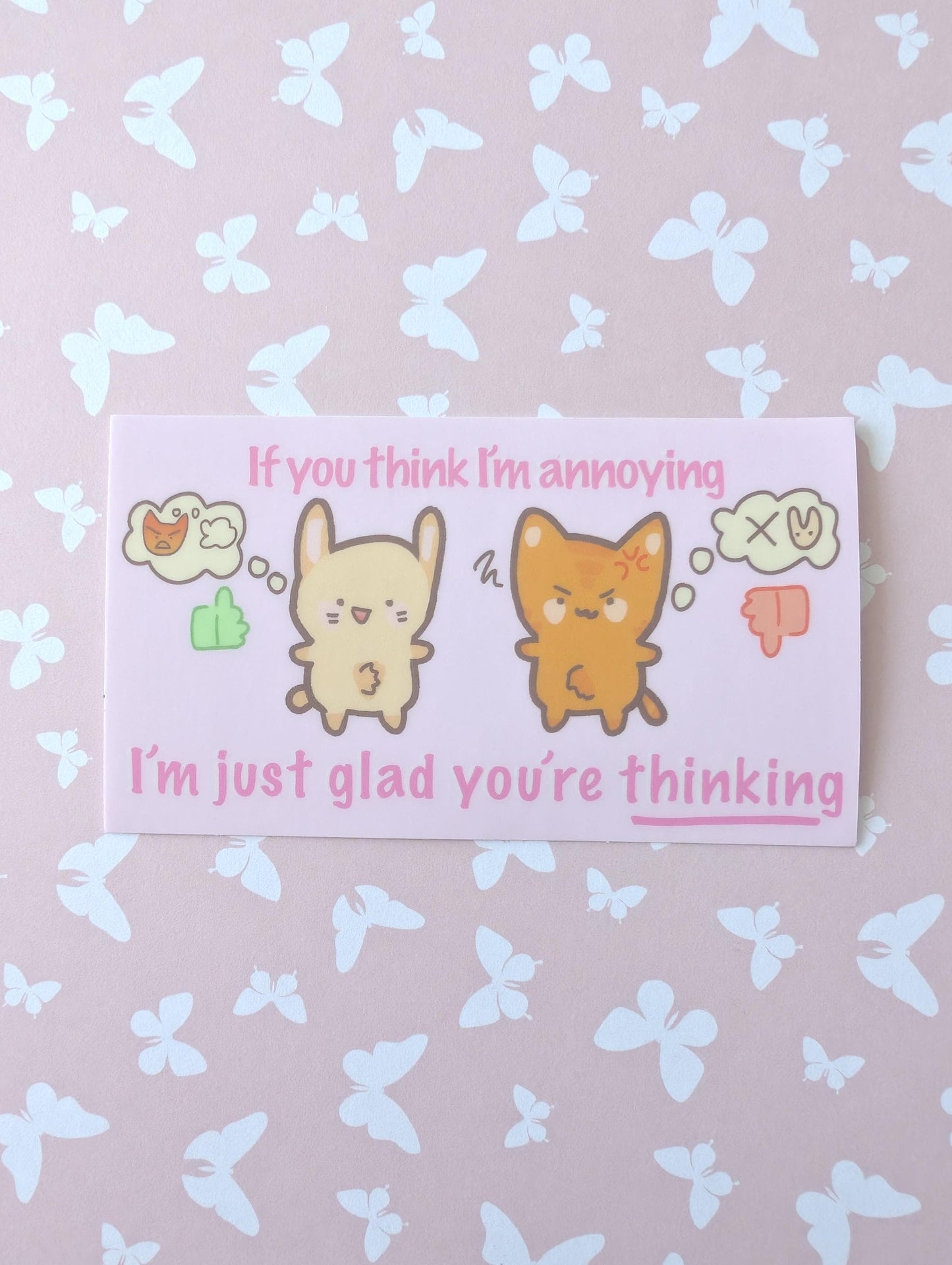 A rectangular printed card with a cute cartoon rabbit and cat, pink text reading 'If you think I'm annoying I'm just glad you're thinking', placed on a pale pink background with a white butterfly pattern.
