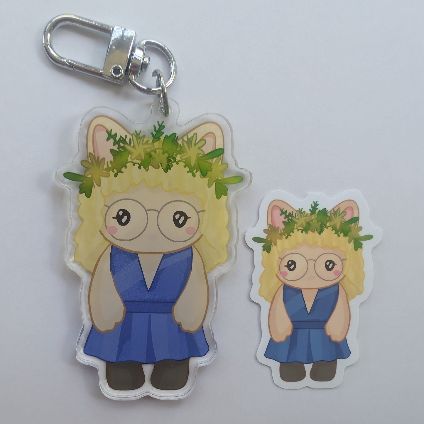 A cartoon-style misty quigley acrylic keychain and matching die-cut sticker featuring a blonde girl with glasses, pink cat-ear ears, and a green leafy crown, shown on a plain light background.
