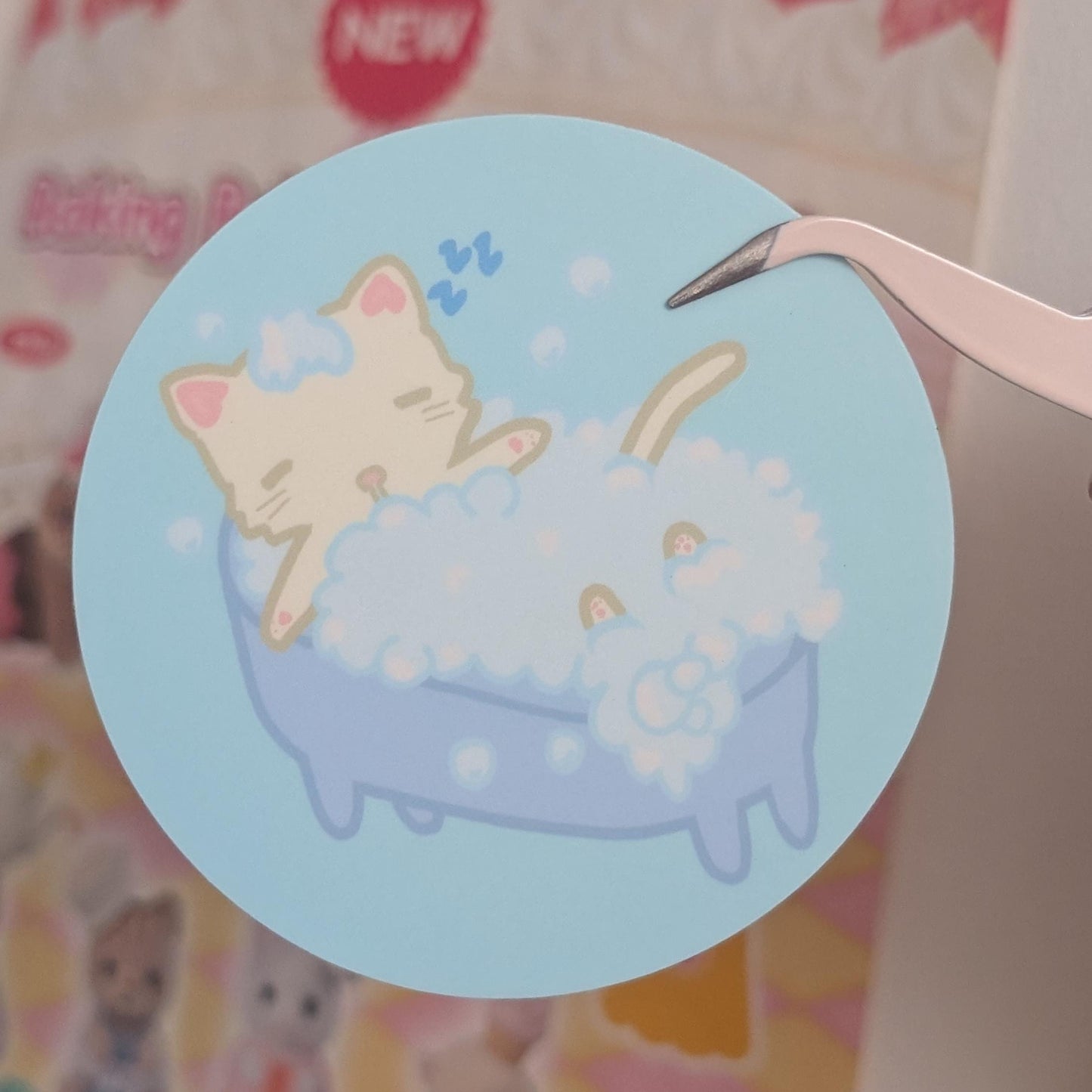 A round sticker featuring a cute illustration of a cream-colored cat sleeping in a light blue bathtub filled with white bubbles, held by tweezers against a blurred pink and white background.