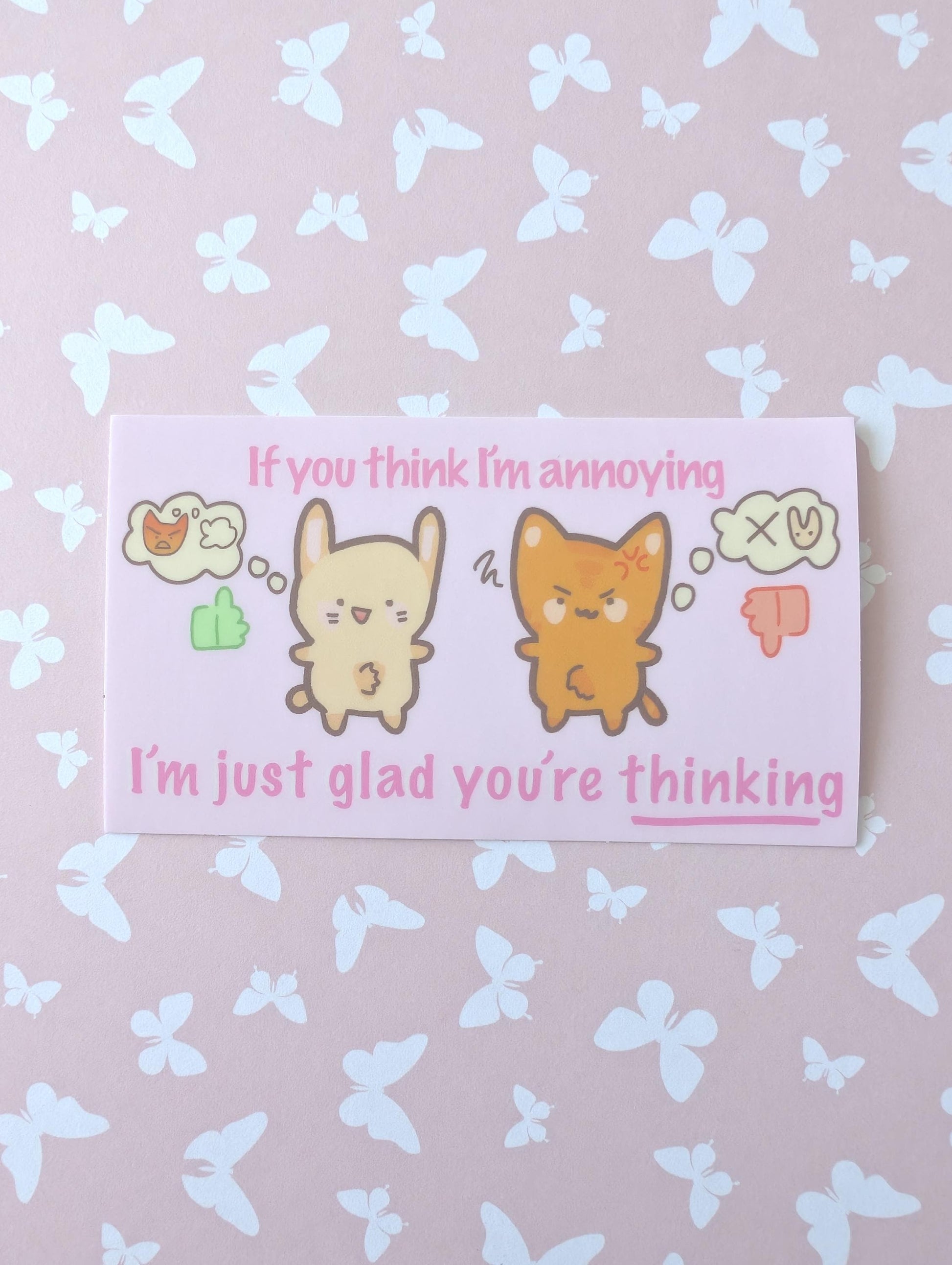 A rectangular printed card with a cute cartoon rabbit and cat, pink text reading 'If you think I'm annoying I'm just glad you're thinking', placed on a pale pink background with a white butterfly pattern.