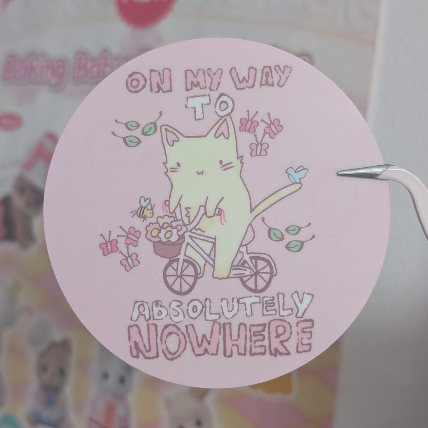 A round pink sticker featuring a cute illustration of a pale green cat riding a bicycle with the text 'ON MY WAY TO ABSOLUTELY NOWHERE', held by tweezers against a blurred pastel background.
