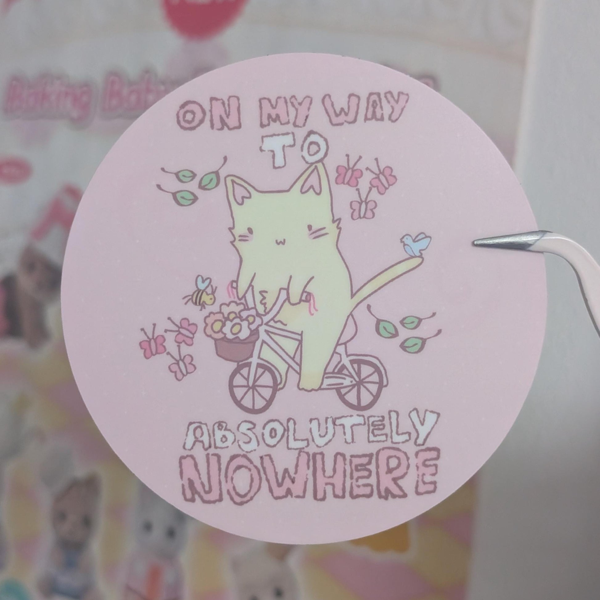 A round pink sticker featuring a cute illustration of a pale green cat riding a bicycle with the text 'ON MY WAY TO ABSOLUTELY NOWHERE', held by tweezers against a blurred pastel background.