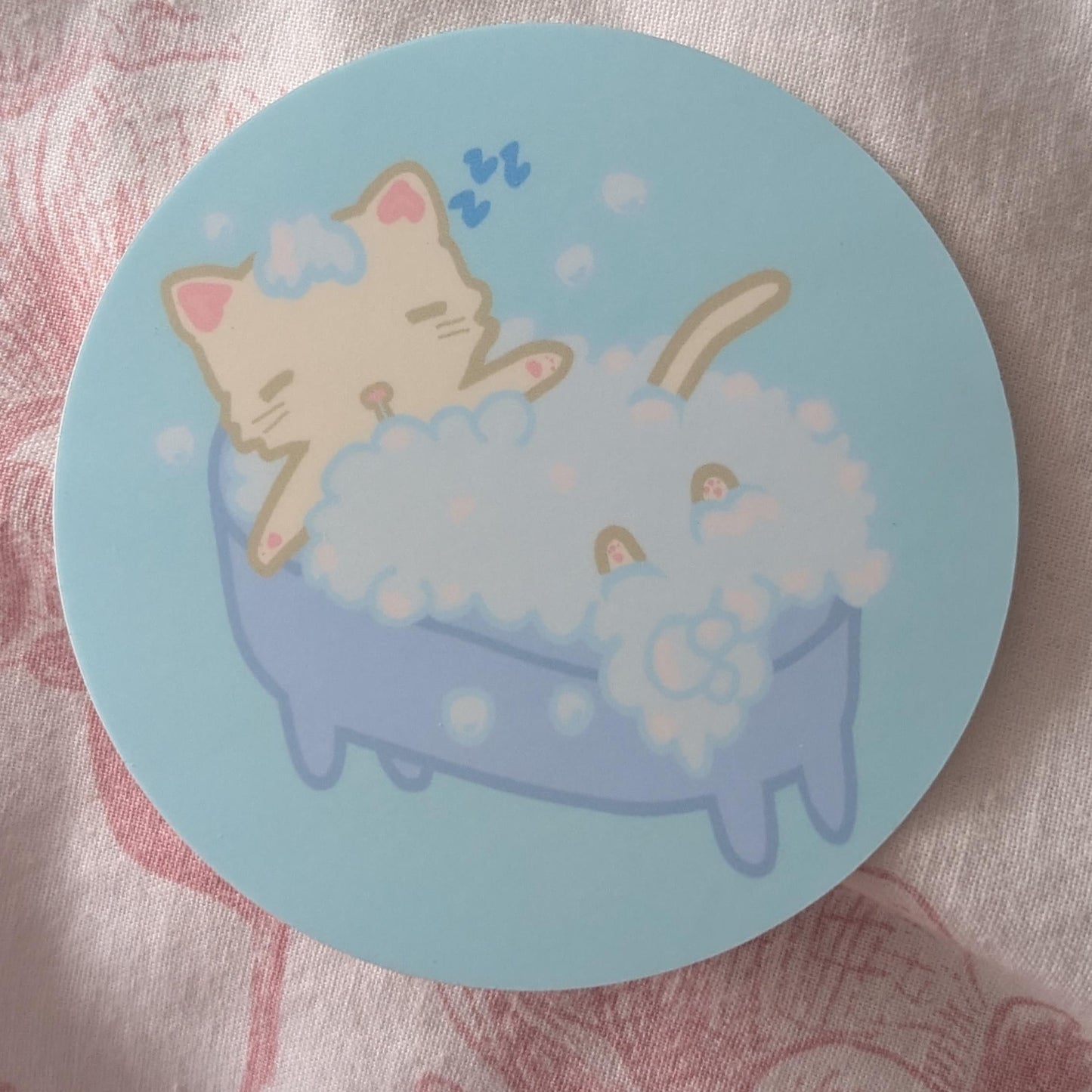 A round sticker or coaster featuring a cute cartoon illustration of a beige cat sleeping in a blue bathtub filled with white bubbles, placed on a pink patterned fabric background.