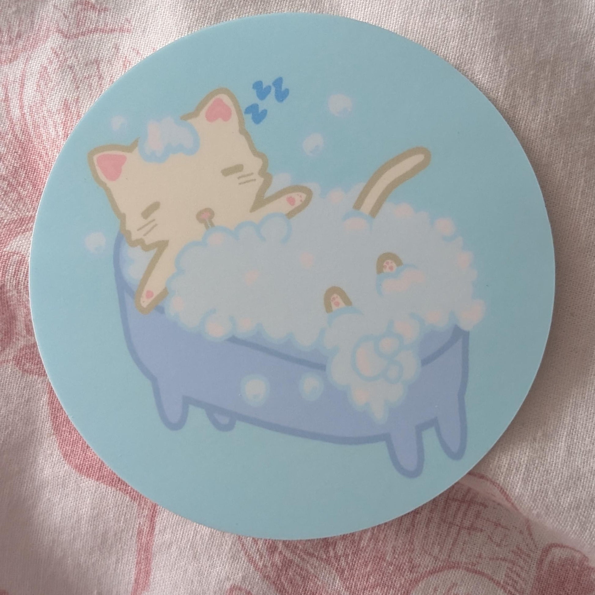 A round sticker or coaster featuring a cute cartoon illustration of a beige cat sleeping in a blue bathtub filled with white bubbles, placed on a pink patterned fabric background.