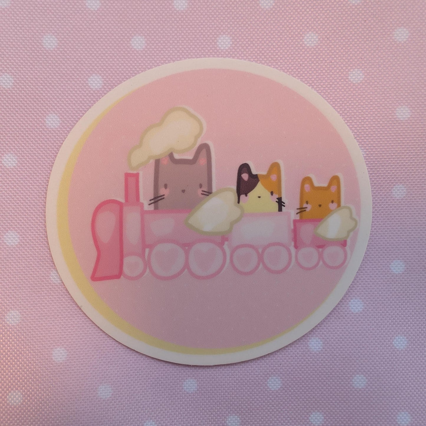 A round sticker featuring a pink train with three cute cartoon cats riding on it, set against a pink polka-dot fabric background.