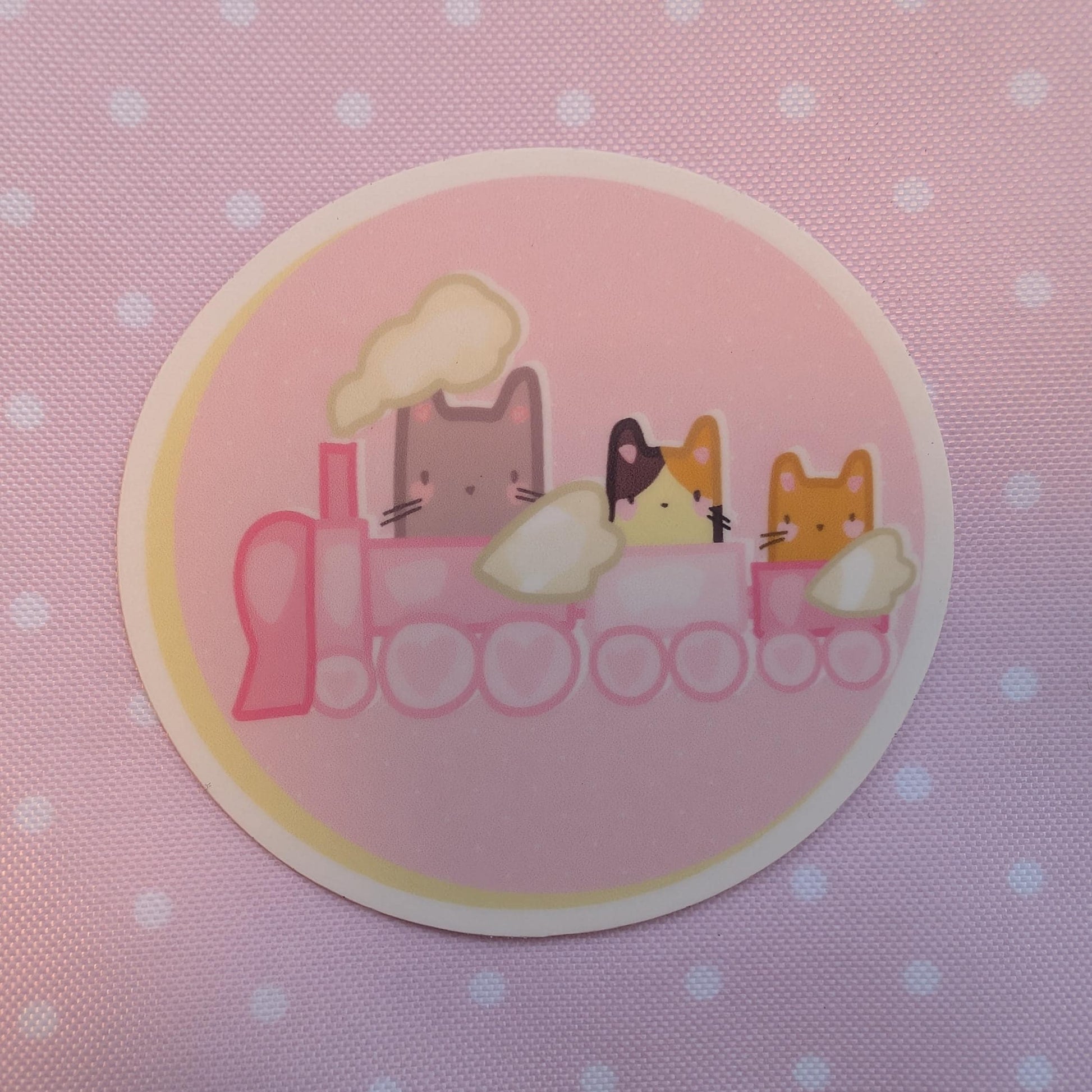 A round sticker featuring a pink train with three cute cartoon cats riding on it, set against a pink polka-dot fabric background.