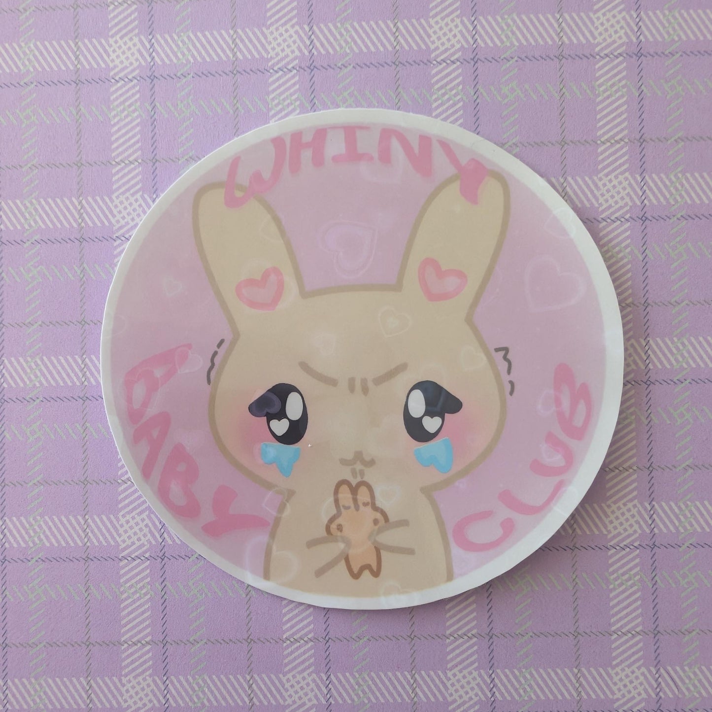 A round white plate or dish featuring a cute cartoon rabbit illustration with pink hearts, blue tears, and the words 'WHINY BABY CLUB' in pink, placed on a lavender plaid surface.