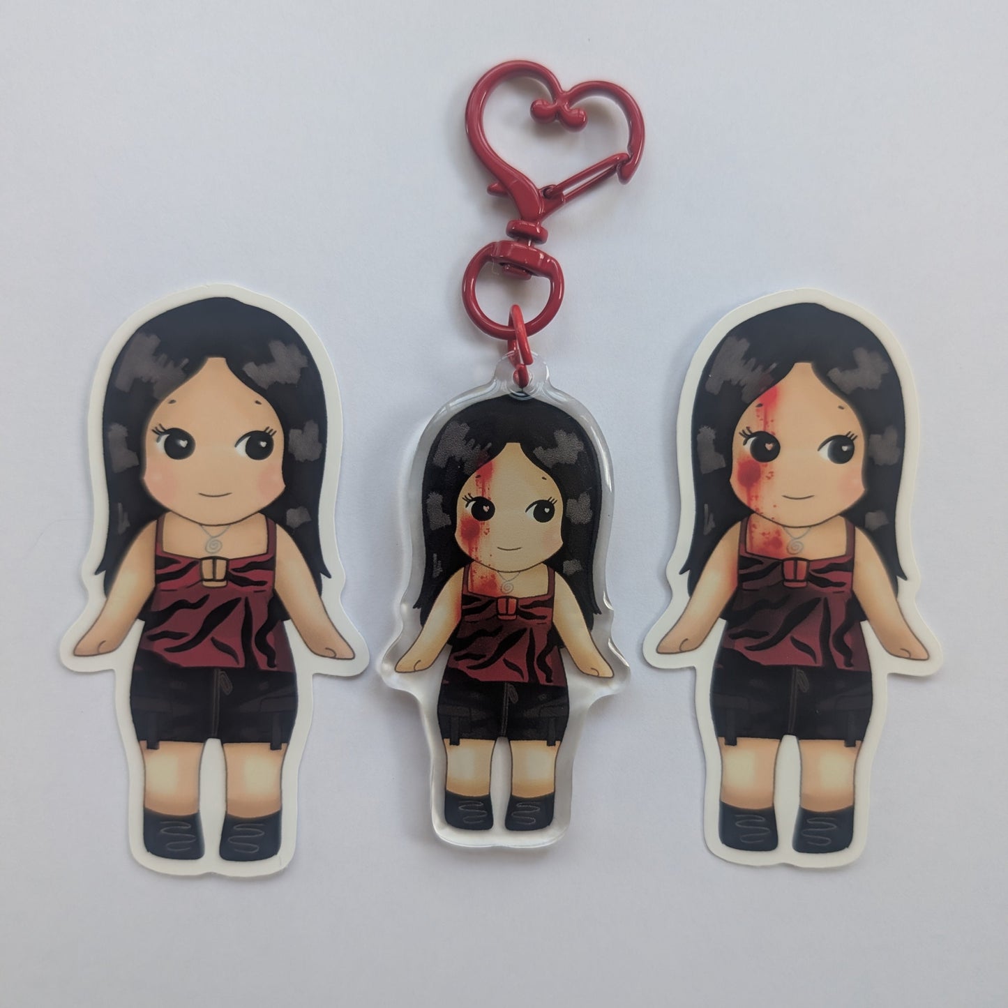 Nikki freeman keychain sonny angel. Three die-cut acrylic keychains featuring a cartoon girl with long black hair, a red sleeveless top, and black shorts, shown on a plain light gray background. The center keychain has a red heart-shaped clasp attached.