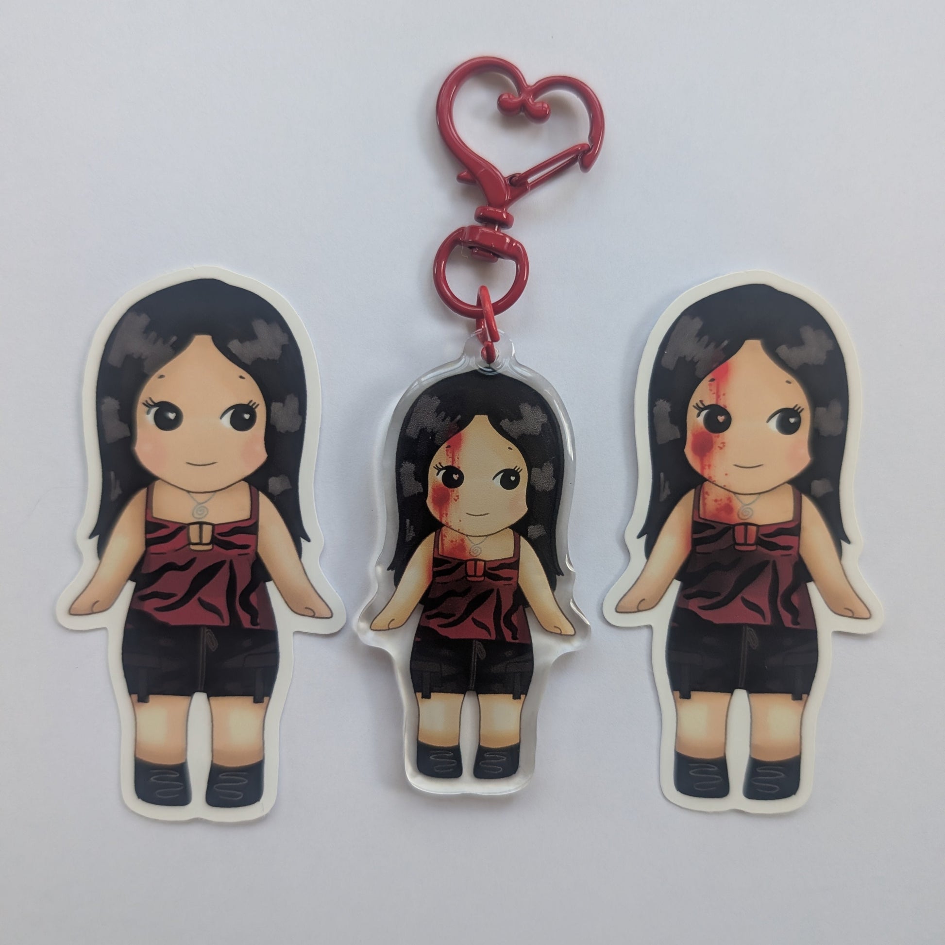 Nikki freeman keychain sonny angel. Three die-cut acrylic keychains featuring a cartoon girl with long black hair, a red sleeveless top, and black shorts, shown on a plain light gray background. The center keychain has a red heart-shaped clasp attached.