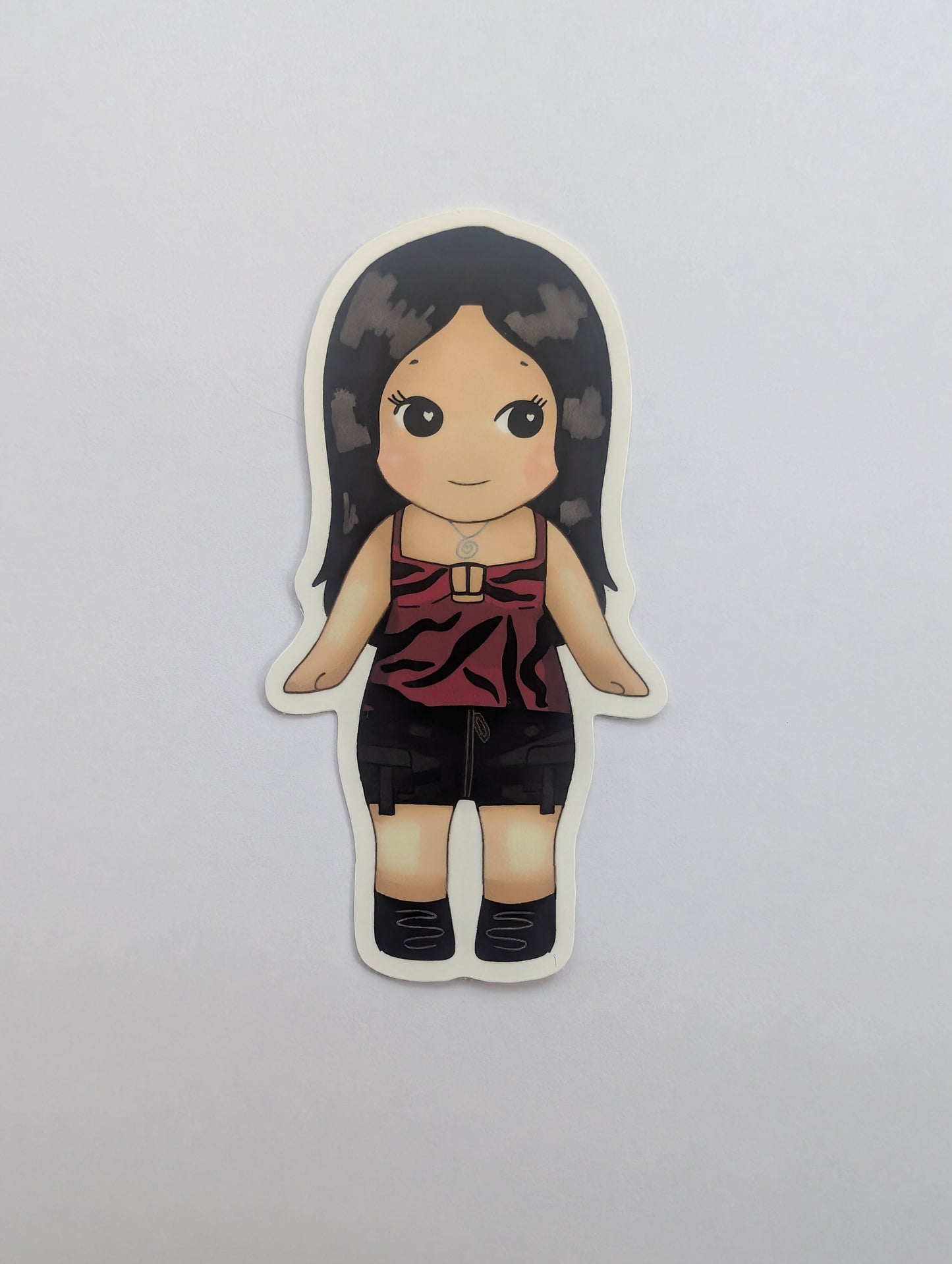 Nikki freeman keychain sonny angel. A die-cut sticker featuring a cartoon-style illustration of a girl with long dark hair, wearing a red and black patterned tank top and black shorts, shown on a plain light gray background.