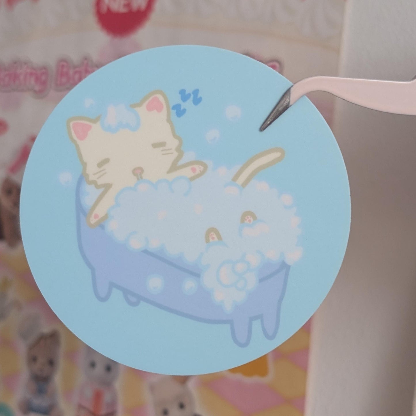 A round sticker featuring a cute illustration of a cream-colored cat and a blue bunny in a bubble bath, held by tweezers against a pastel-themed background.