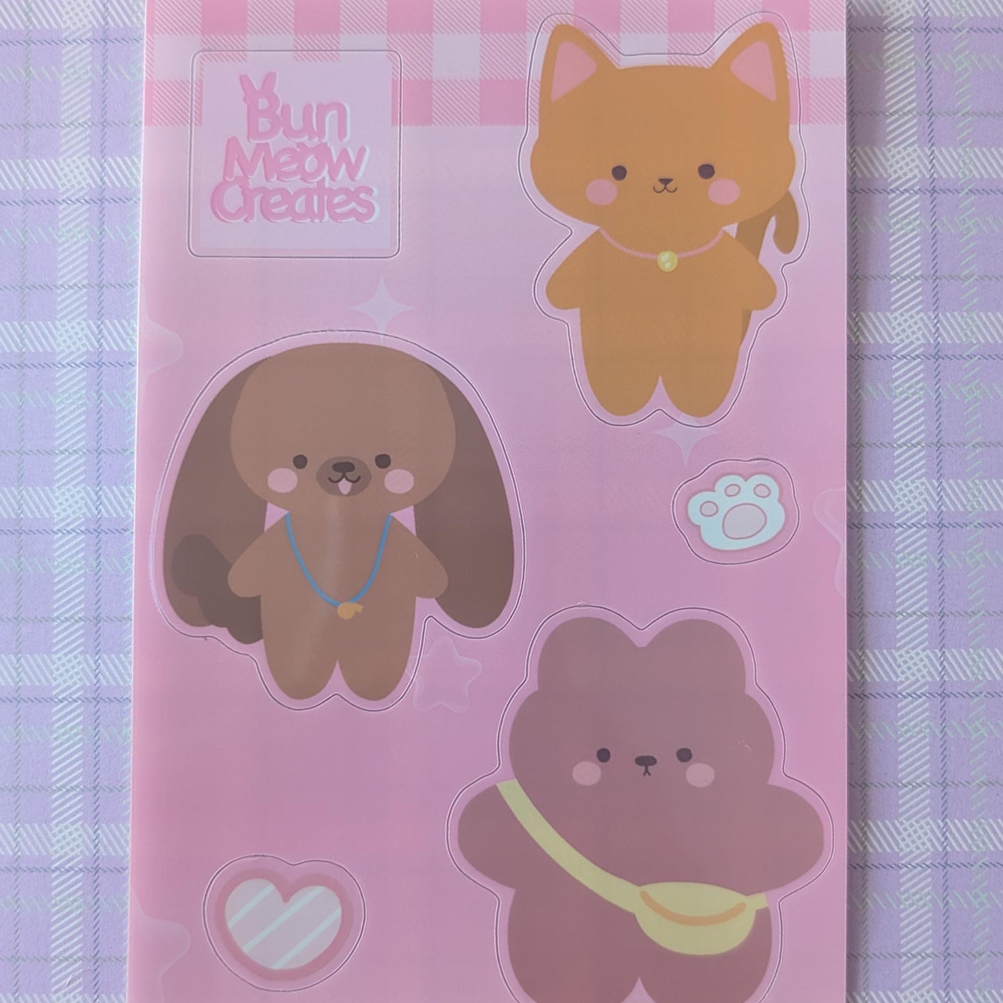 A pink sticker sheet featuring cute cartoon animal characters, including a ginger cat, a brown dog, and a pink bear, with the brand name 'Bun Mett Creates' printed in the top left corner, displayed on a purple and white checkered fabric background.