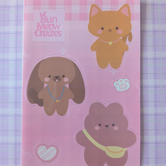 A pink sticker sheet featuring cute cartoon animal characters, including a ginger cat, a brown dog, and a pink bear, with the brand name 'Bun Mett Creates' printed in the top left corner, displayed on a purple and white checkered fabric background.