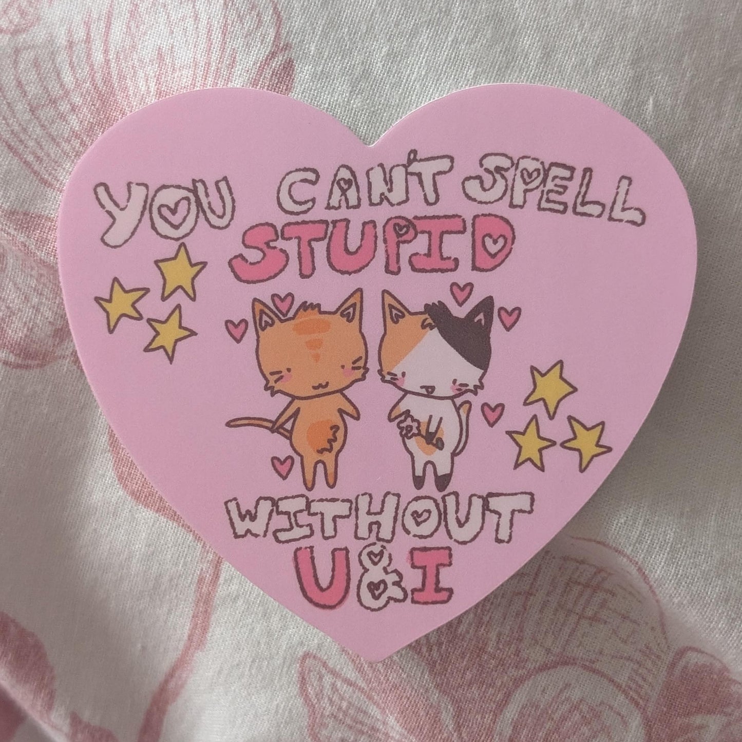 A pink heart-shaped sticker featuring the text 'YOU CAN'T SPELL STUPID WITHOUT U&I' in stylized lettering, decorated with cartoon cats, hearts, and stars, placed on a pink and white patterned fabric background.
