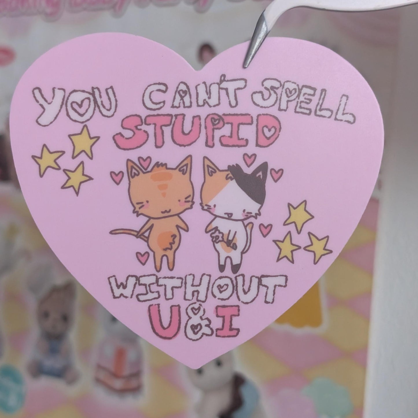 A pink heart-shaped paper tag with a cute cartoon cat illustration and the text 'YOU CAN'T SPELL STUPID WITHOUT U&I', held by a white plastic hook on a blurred pastel-themed background.
