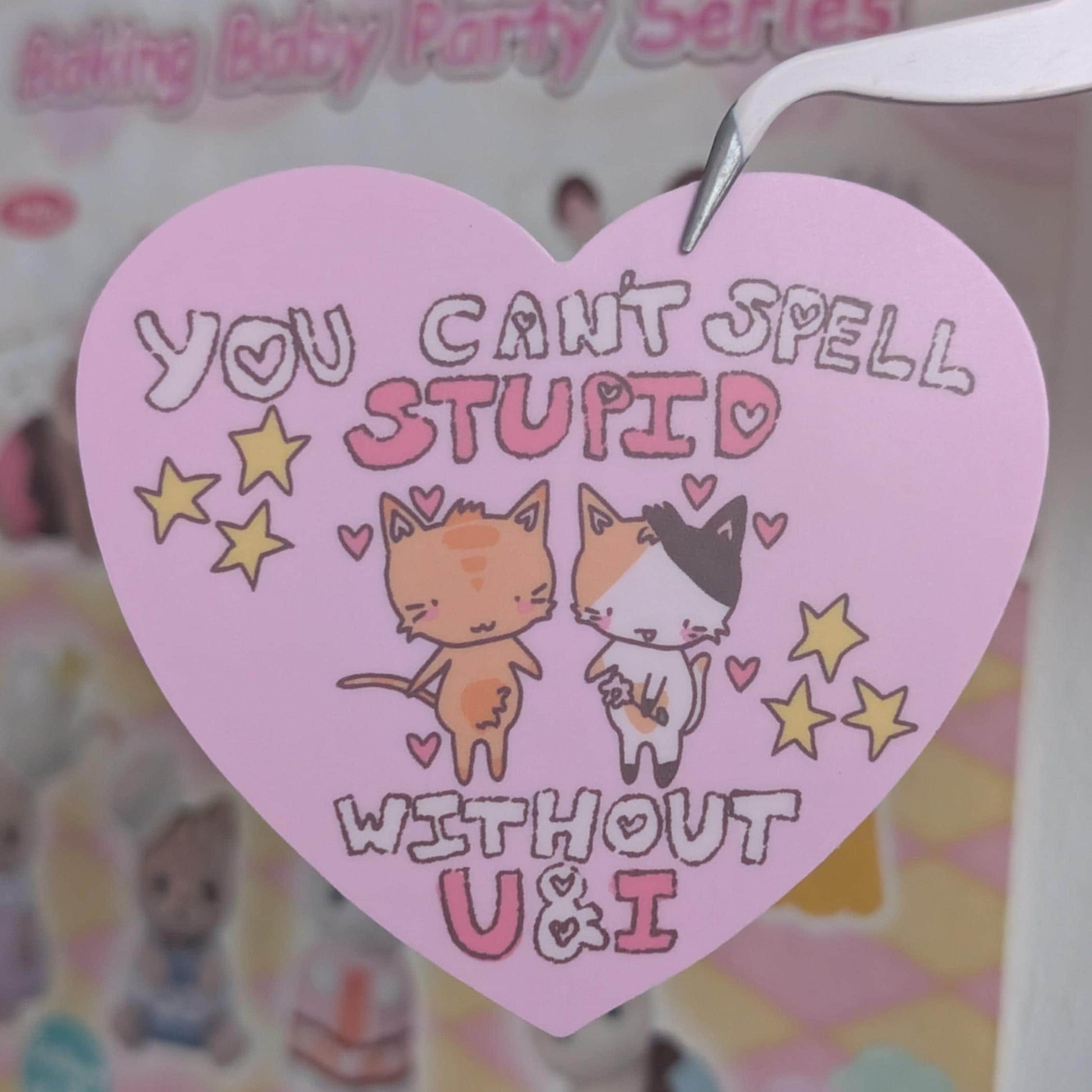 A pink heart-shaped paper tag with a metal loop at the top, featuring the text 'YOU CAN'T SPELL STUPID WITHOUT U&I' and cute cartoon cats, held by tweezers against a blurred pastel background.