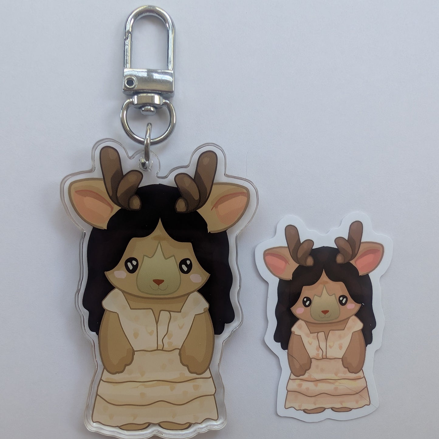 Two cartoon lottie matthews deer-themed keychain charms or stickers on a plain light background, one larger and one smaller, both with antlers, dark hair, and a cream dress, plus a silver key ring and clasp on the larger piece.