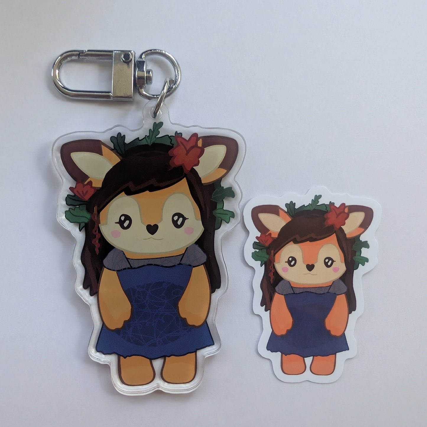 Two cartoon deer shauna shipman character acrylic keychain charms with a metal clasp, shown on a plain light background. One is larger and one is smaller, both featuring brown hair, a blue dress, orange face and legs, and a green floral crown.
