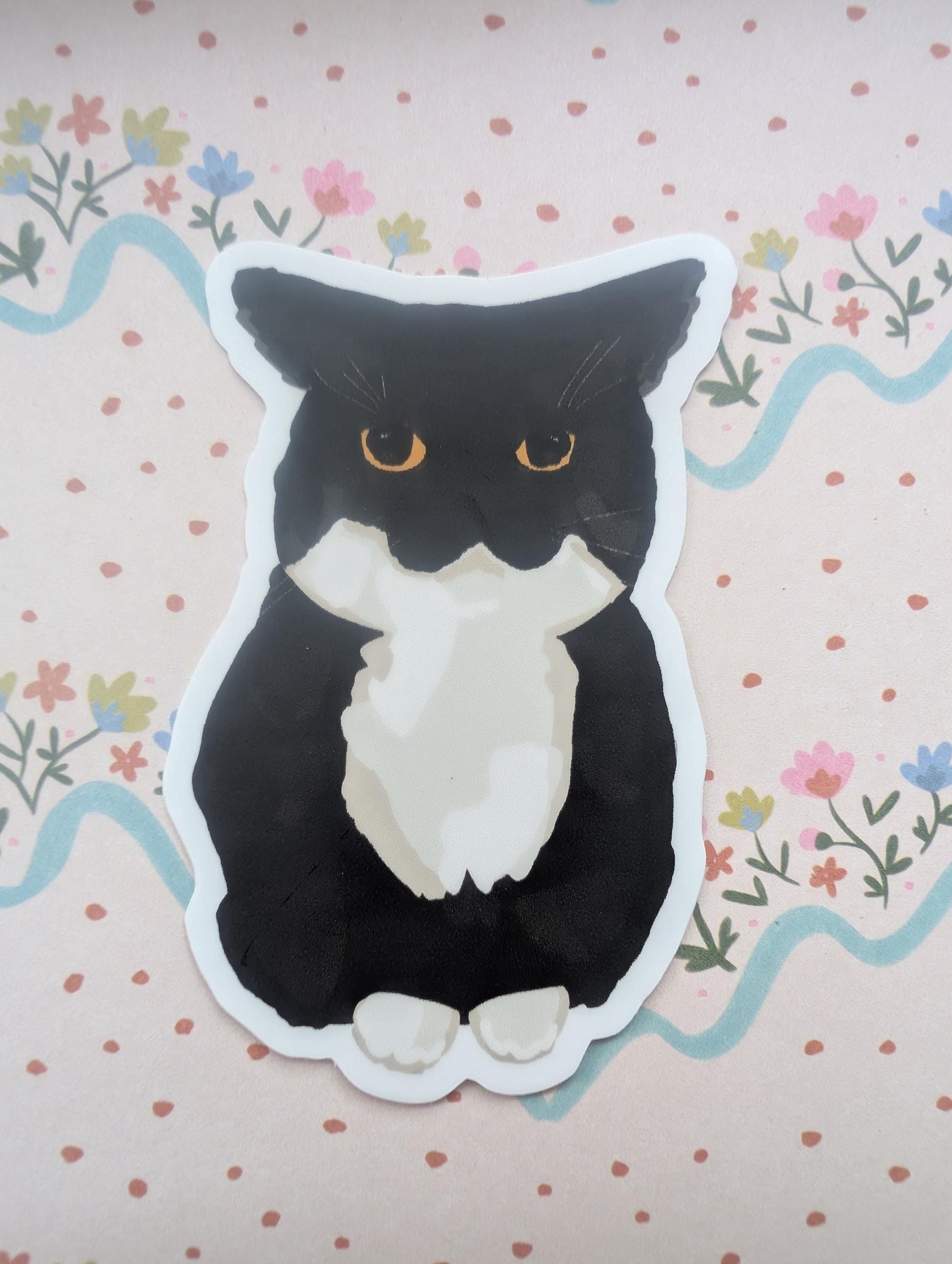 A die-cut sticker of a black and white cat with orange eyes, placed on a pink floral background.