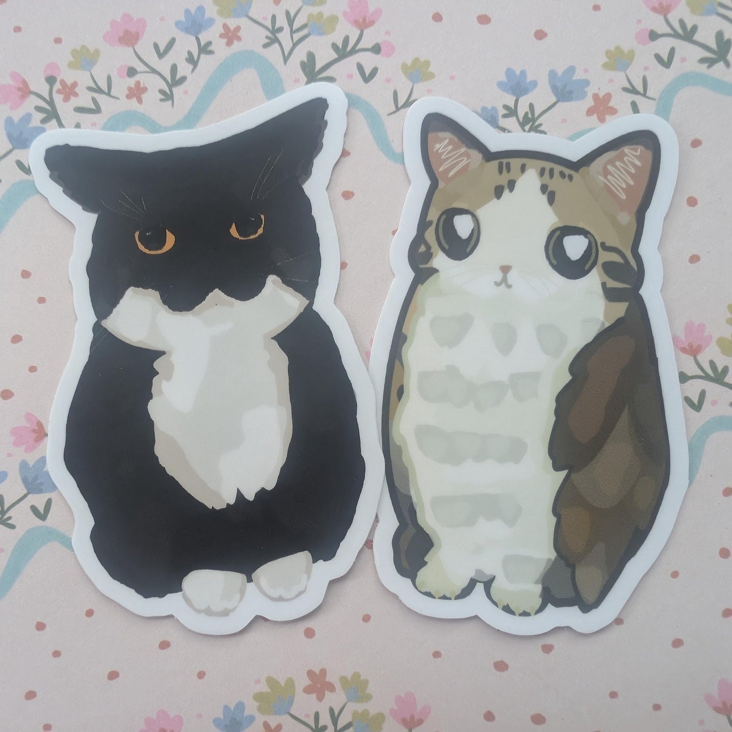 Two die-cut stickers featuring cute illustrated cats, one black and white and one brown and cream, placed on a pink floral background.
