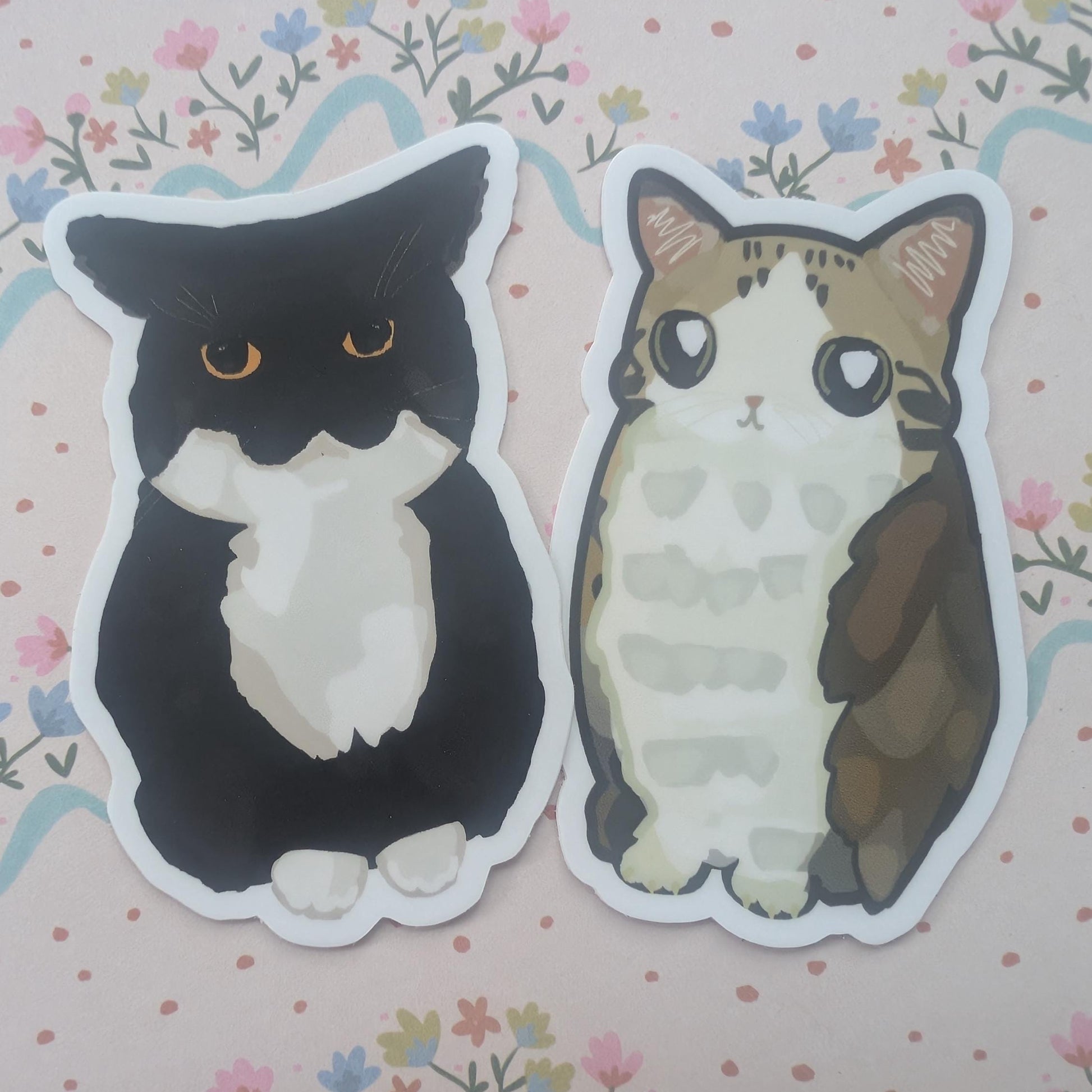 Two die-cut stickers featuring cute illustrated cats, one black and white and one brown and cream, placed on a pink floral background.