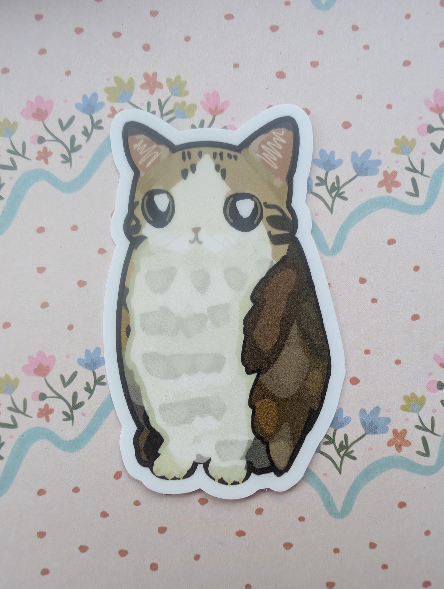 A die-cut sticker featuring a cute illustrated tabby and white cat with large eyes, placed on a pink floral background.